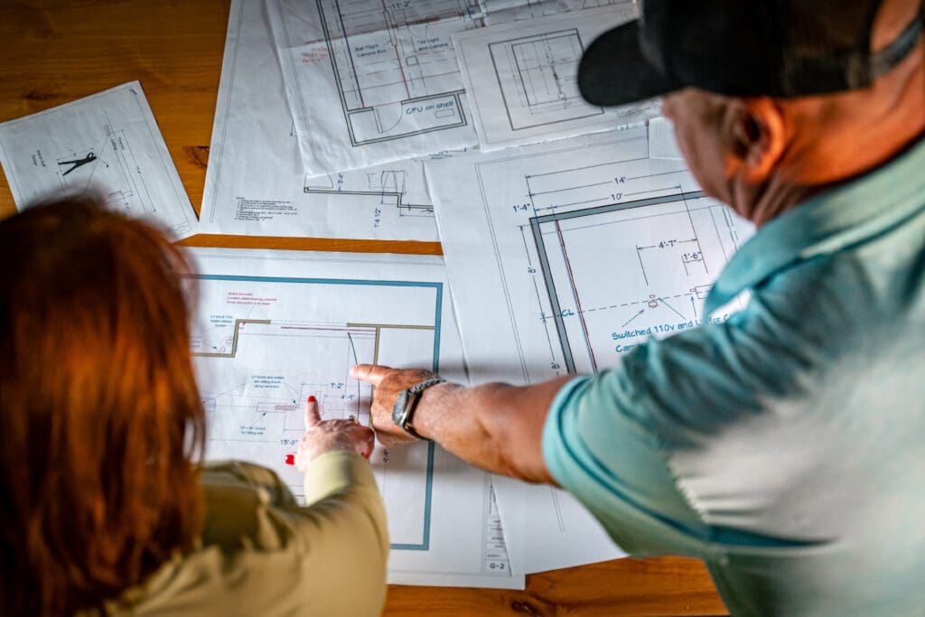 Two people are engrossed in studying blueprints on a table while occasionally glancing at their state-of-the-art golf simulator nearby.